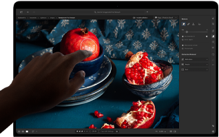 MacBook Pro พร้อมจอภาพ PROLED XDR แสดงภาพนิ่งของผลทับทิมพร้อมเซรามิกและสิ่งทอสีฟ้า มือหนึ่งยื่นออกไปที่หน้าจอเพื่อโต้ตอบกับภาพ